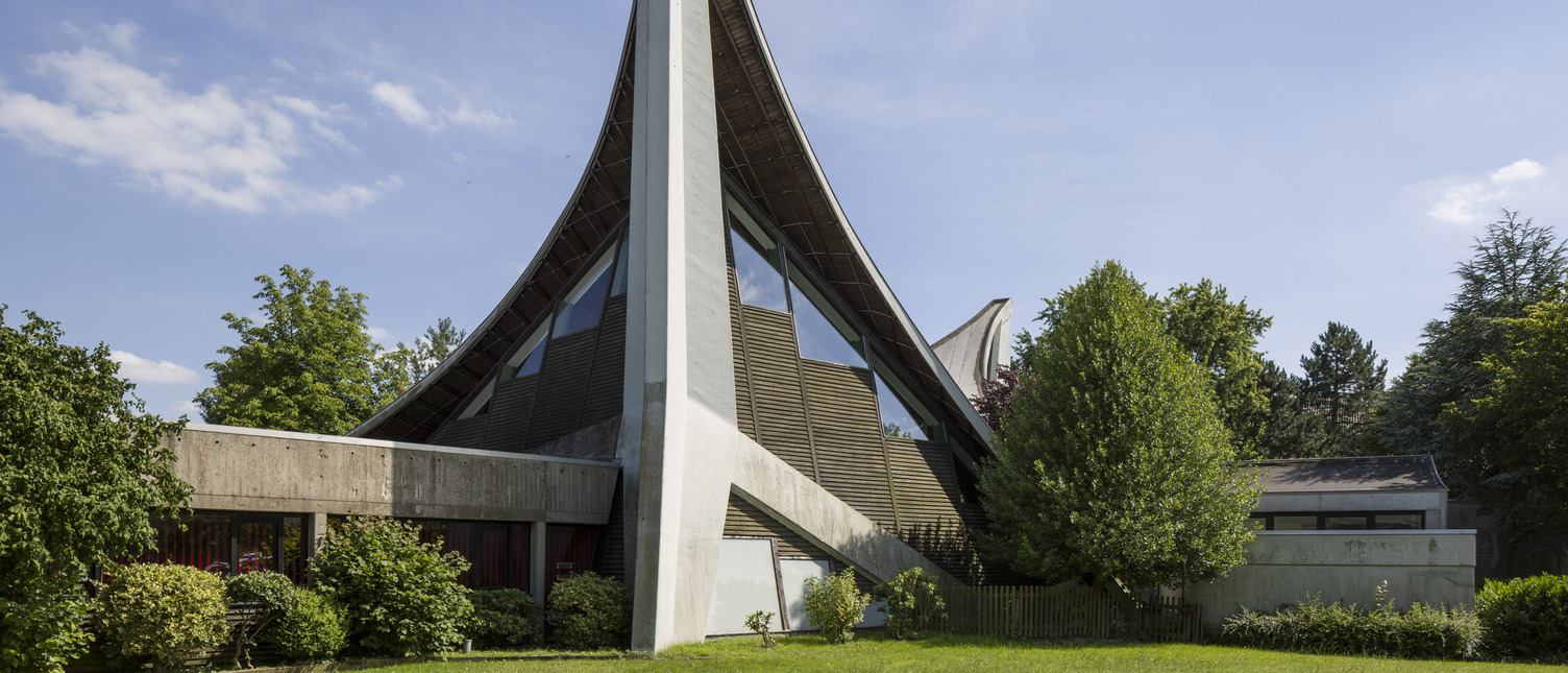 Das Bild zeigt ein modernes Gebäude mit einer charakteristischen geschwungenen Dachkonstruktion und einer hohen, schrägen Wand. Umgeben von Bäumen und Sträuchern, steht das Gebäude in einer grünen Umgebung unter einem klaren blauen Himmel.
