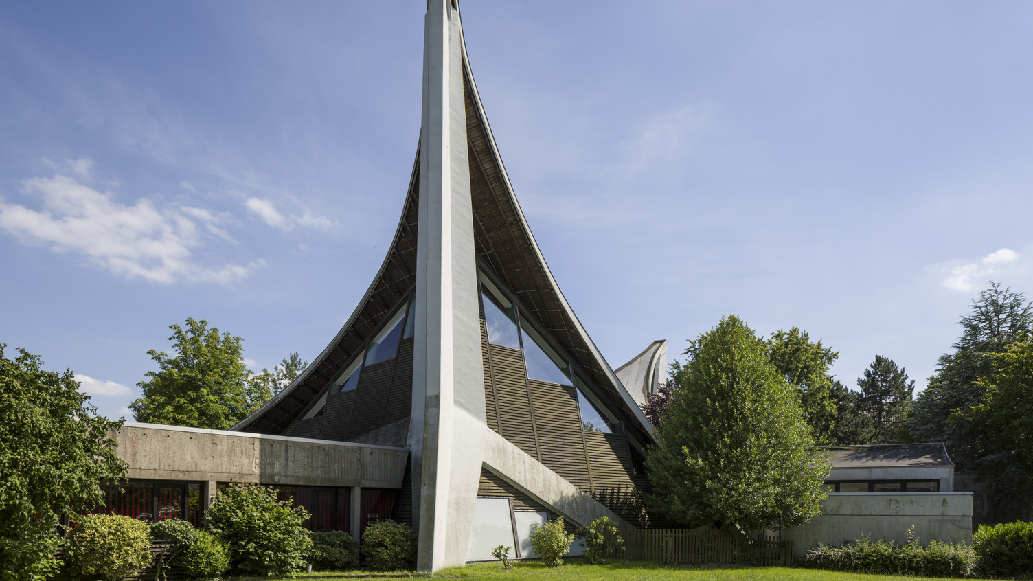 Das Bild zeigt ein modernes Gebäude mit einer charakteristischen geschwungenen Dachkonstruktion und einer hohen, schrägen Wand. Umgeben von Bäumen und Sträuchern, steht das Gebäude in einer grünen Umgebung unter einem klaren blauen Himmel.
