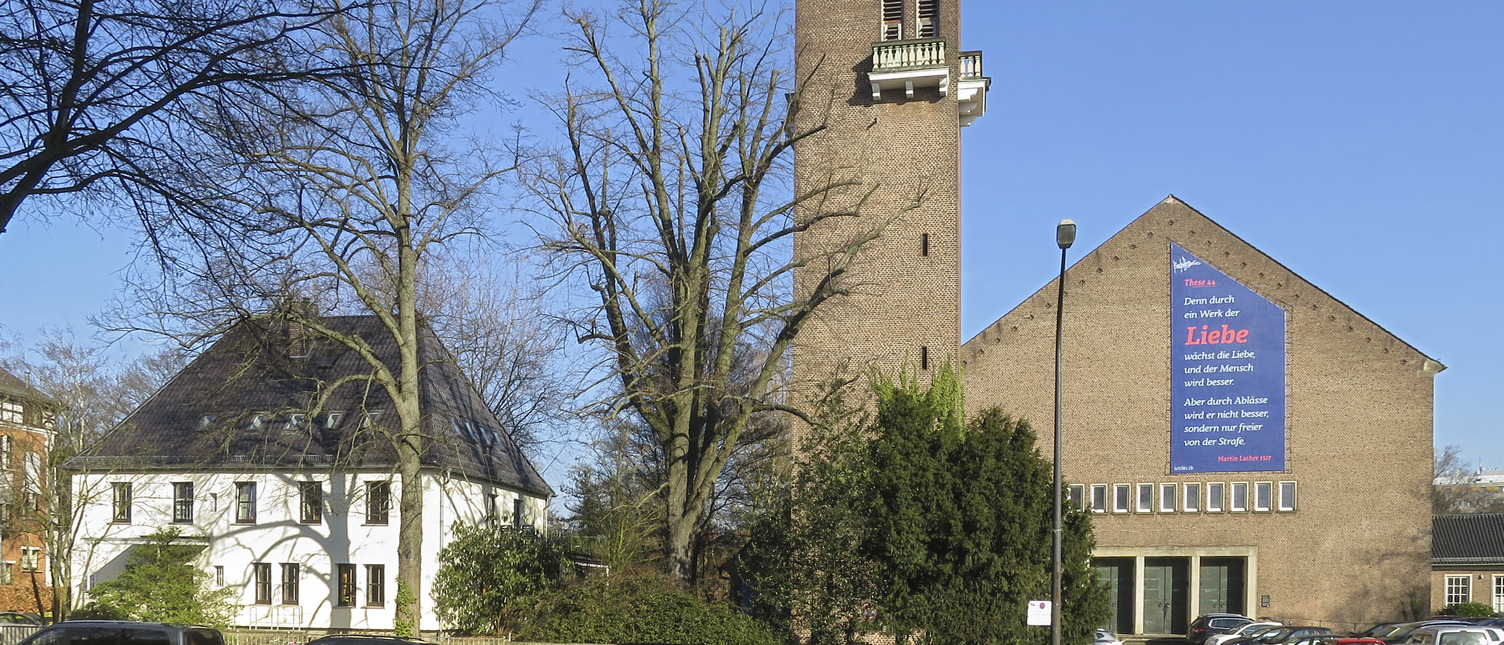 Das Bild zeigt eine Kirche mit einem hohen Turm und einem großen, bunten Banner, das eine Botschaft über Liebe enthält. Im Vordergrund stehen mehrere Autos, und im Hintergrund sind Bäume und ein weiteres Gebäude zu sehen. Der Himmel ist blau und klar.