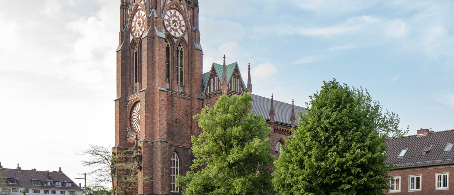 Ein historischer Glockenturm aus Backstein steht im Fokus des Bildes, umgeben von Bäumen. Der Turm hat auffällige Ziffernblätter und eine reich verzierte Fassade. Im Hintergrund sind weitere Gebäude und der Himmel sichtbar, der teilweise bewölkt ist.