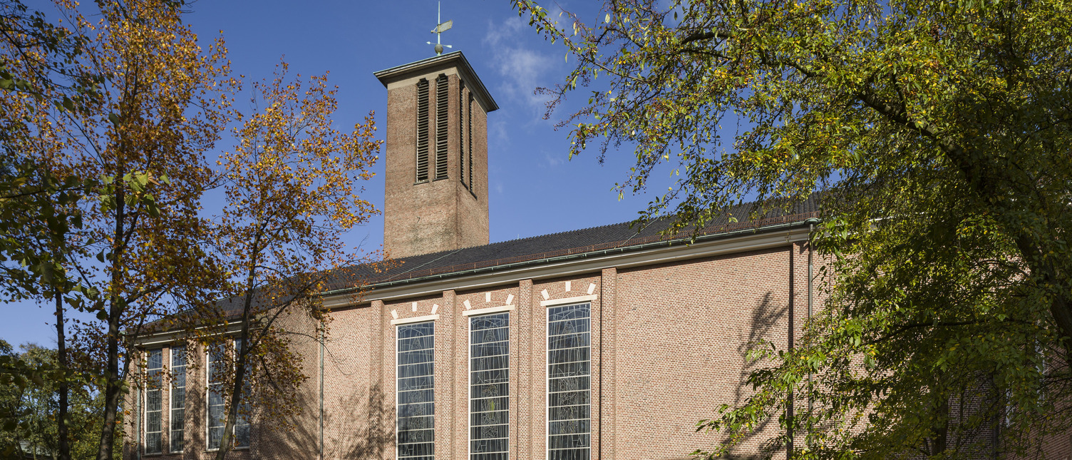 Das Bild zeigt eine Backsteinkirche mit großen Fenstern und einem schmalen Turm. Die Fassade ist von Bäumen umgeben. Ein grünes Eingangstor ist gut sichtbar, und der Himmel ist blau mit wenigen Wolken.