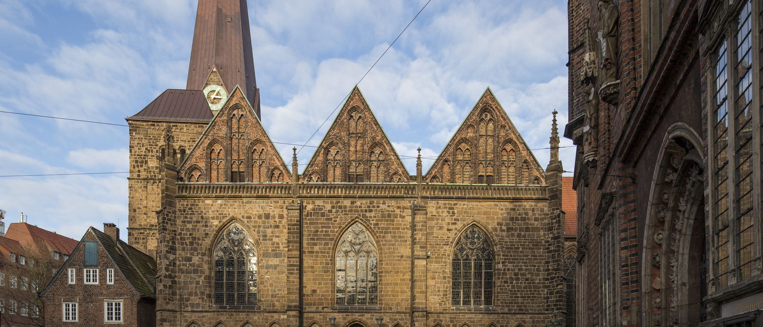 Ein Blick auf die historische Kirche mit einem hohen, spitzen Turm und markanten Ziegelsteinfassaden. Vor dem Gebäude ist ein Platz erkennbar, der von anderen Bauwerken umgeben ist. Der Himmel ist blau mit einigen Wolken.