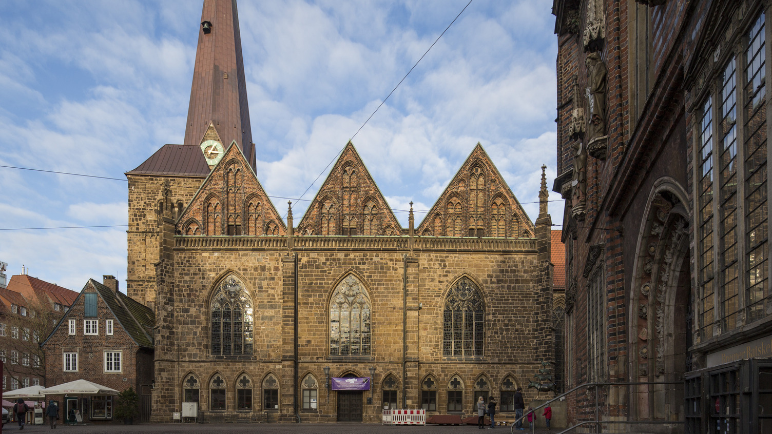 Ein Blick auf die historische Kirche mit einem hohen, spitzen Turm und markanten Ziegelsteinfassaden. Vor dem Gebäude ist ein Platz erkennbar, der von anderen Bauwerken umgeben ist. Der Himmel ist blau mit einigen Wolken.