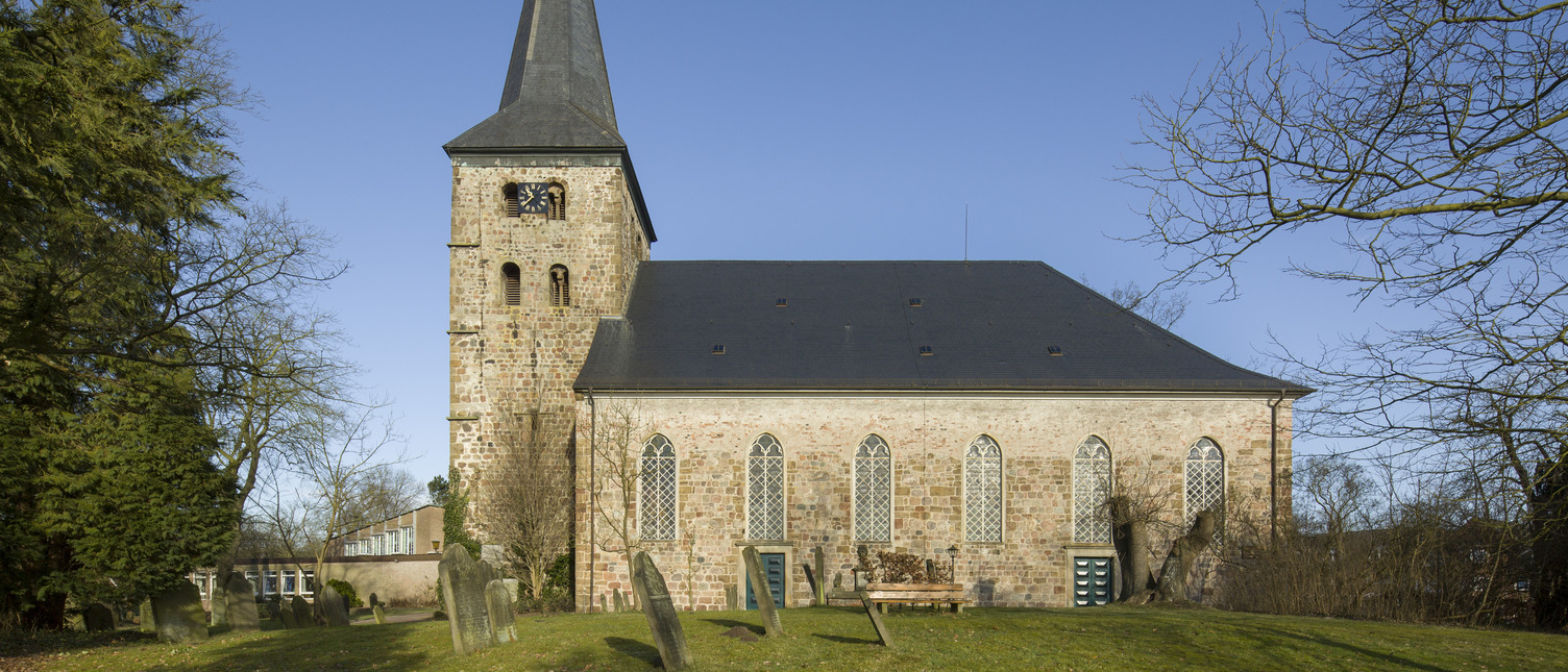 Die Bildbeschreibung zeigt eine Kirche mit einem hohen, spitzen Turm und charakteristischen Fenstern. Der Gebäudekörper ist aus helleren Ziegelsteinen, umgeben von einem ruhigen, grünen Rasen und einigen Grabsteinen im Vordergrund. Der Himmel ist klar und blau.