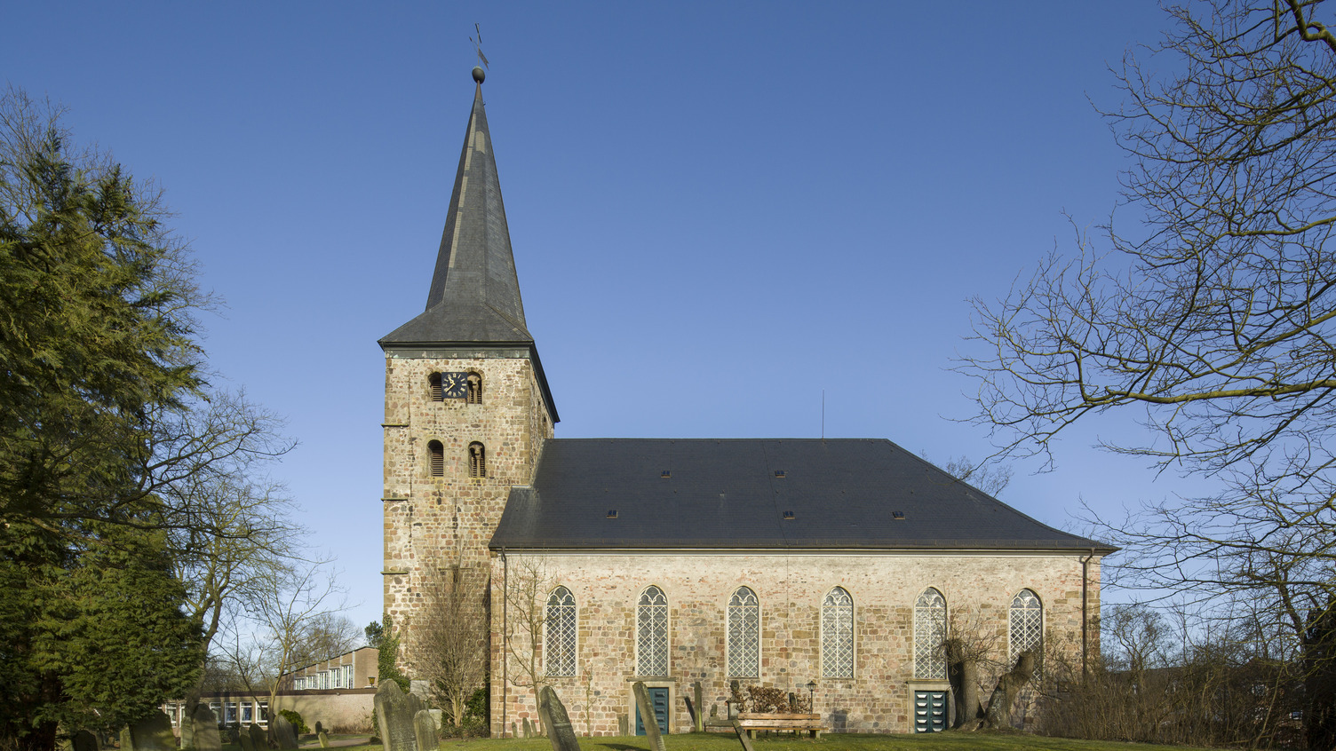 Die Bildbeschreibung zeigt eine Kirche mit einem hohen, spitzen Turm und charakteristischen Fenstern. Der Gebäudekörper ist aus helleren Ziegelsteinen, umgeben von einem ruhigen, grünen Rasen und einigen Grabsteinen im Vordergrund. Der Himmel ist klar und blau.