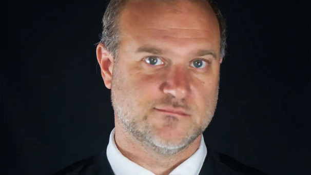 Ein Mann mit kurzen, grauen Haaren und einem leichten Bart schaut direkt in die Kamera. Er trägt einen dunklen Anzug und ein weißes Hemd. Der Hintergrund ist schwarz, was sein Gesicht hervorhebt. Sein Ausdruck wirkt ernst.