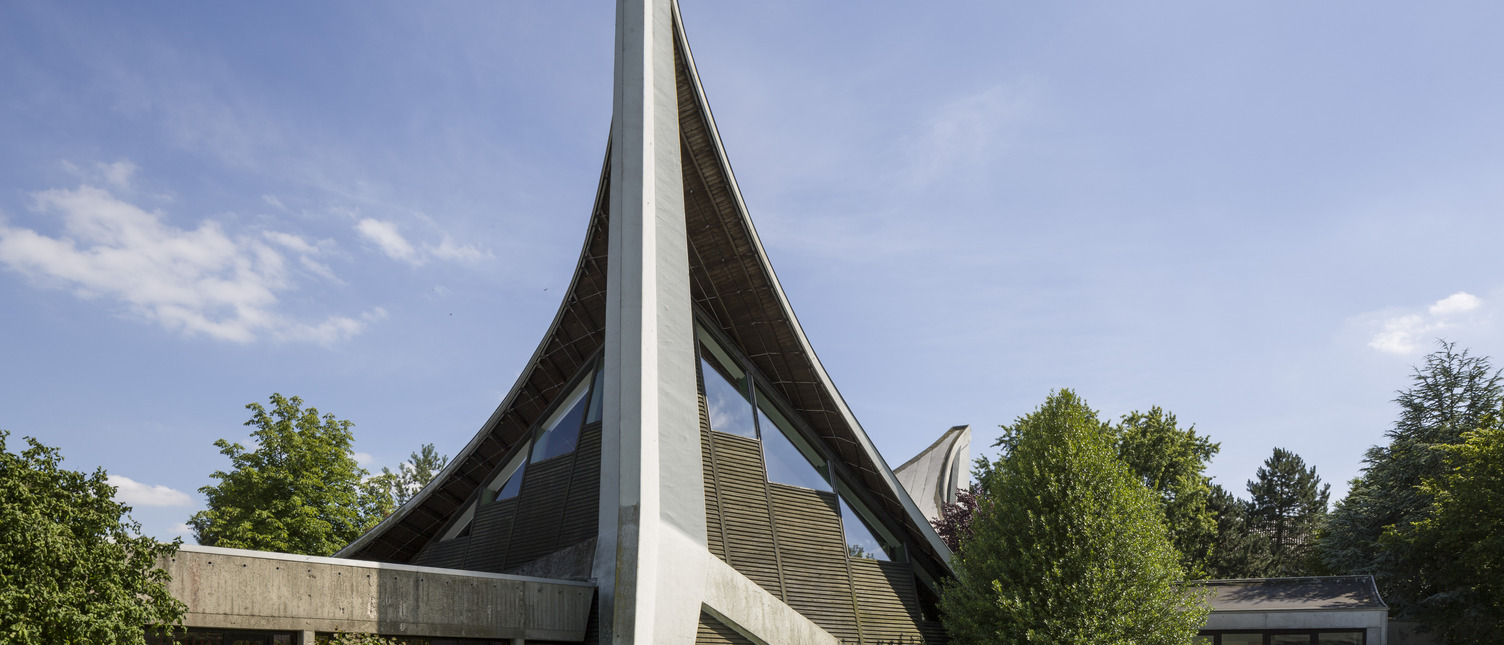 Das Bild zeigt ein modernes Gebäude mit einer charakteristischen geschwungenen Dachkonstruktion und einer hohen, schrägen Wand. Umgeben von Bäumen und Sträuchern, steht das Gebäude in einer grünen Umgebung unter einem klaren blauen Himmel.