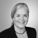 Porträt einer lächelnden Frau mit grauen Haaren in einem schwarzen Blazer. Sie hat eine helle Bluse und trägt eine Halskette. Der Hintergrund ist in sanften Grautönen gehalten.