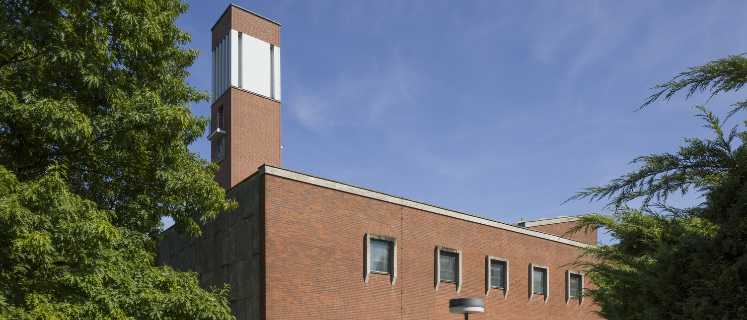 Ein modernes, zweigeschossiges Ziegelgebäude mit großen Fenstern und einem markanten, höheren Turm. Der Eingangsbereich ist durch ein transparentes Vordach geschützt. Um das Gebäude herum sind grüne Bäume und Sträucher sichtbar, bei klarem Himmel und Sonnenschein.