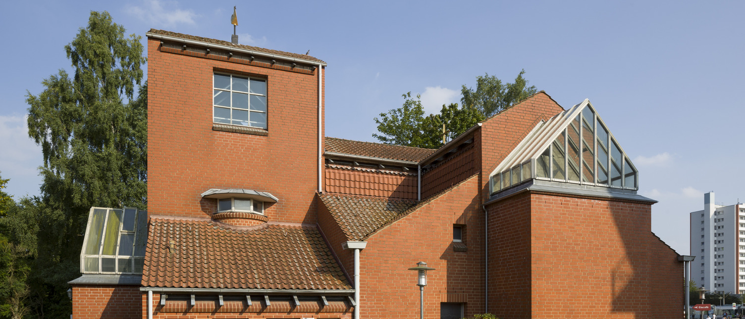 Ein modernes, zweigeschossiges Gebäude aus roten Ziegeln. Es hat ein auffälliges, geneigtes Dach mit einer Glasstruktur und große Fenster. Vor dem Gebäude befindet sich eine gepflegte Hecke und ein kleiner Weg. Im Hintergrund sind Bäume und ein urbanes Umfeld sichtbar.