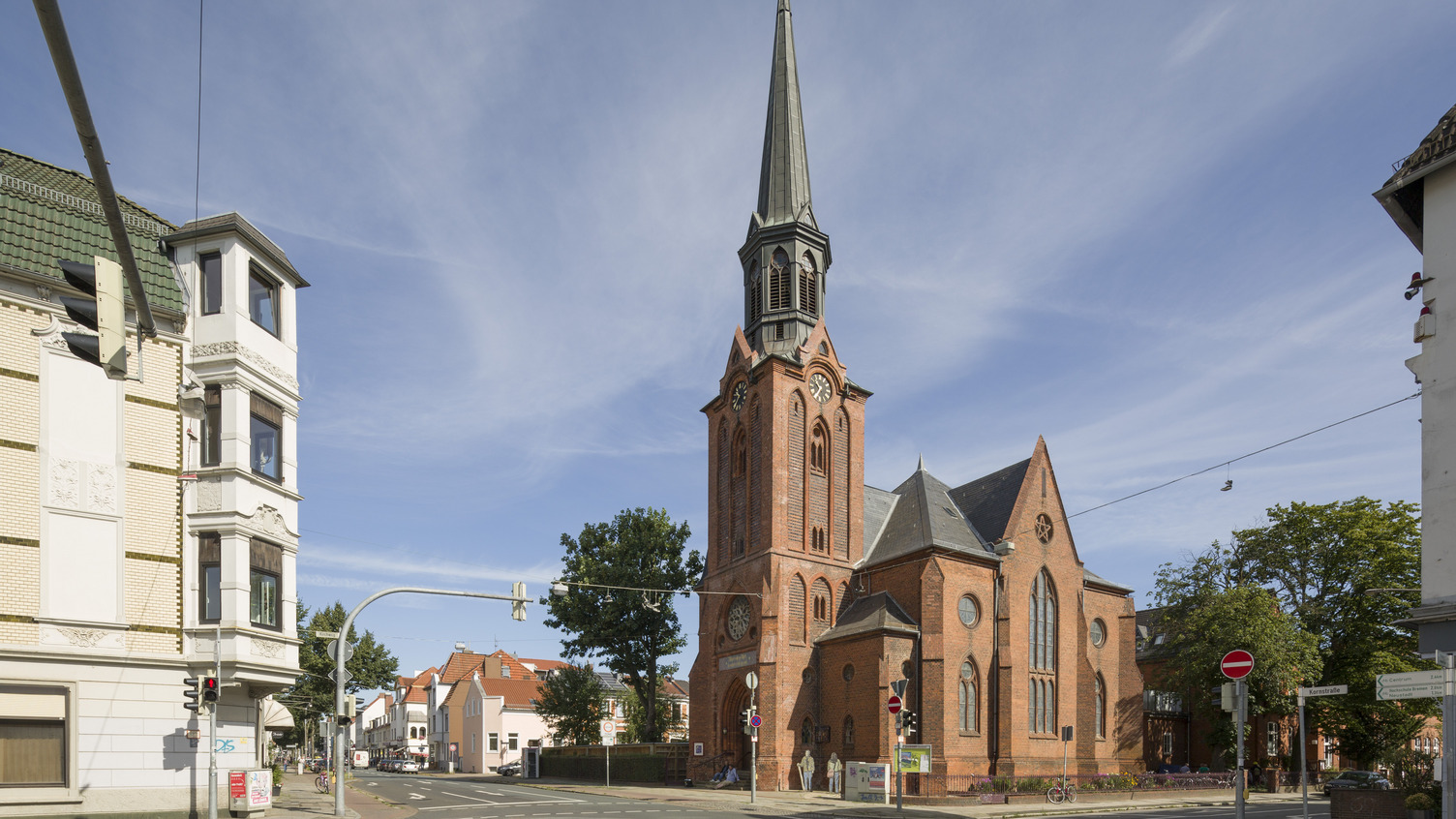 Ein historisches, rotes Backsteingebäude mit einem hohen, spitzen Turm und Zifferblättern an der Fassade. Es steht an einer Straßenecke, umgeben von Wohngebäuden und Bäumen unter einem klaren blauen Himmel. Verkehrsampeln und Straßenschilder sind ebenfalls sichtbar.
