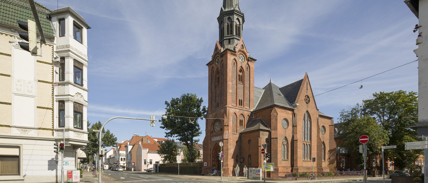 Ein historisches, rotes Backsteingebäude mit einem hohen, spitzen Turm und Zifferblättern an der Fassade. Es steht an einer Straßenecke, umgeben von Wohngebäuden und Bäumen unter einem klaren blauen Himmel. Verkehrsampeln und Straßenschilder sind ebenfalls sichtbar.