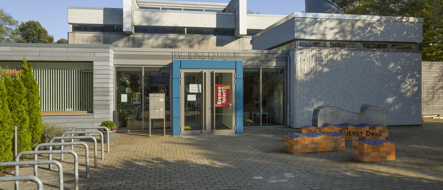 Das Bild zeigt das moderne Gebäude eines Gemeindezentrums mit einer blauen Eingangstür. Vor dem Gebäude befindet sich ein gepflasterter Platz mit Fahrradbügeln und einem bunten Sitzbereich aus Steinen. Die Umgebung ist hell und sonnig, mit Bäumen im Hintergrund.