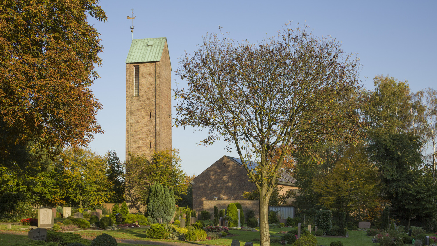 Ein Blick auf einen Friedhof mit sorgfältig gepflegten Gräbern und bunten Blumenbeeten. Im Hintergrund steht ein hoher, rechteckiger Kirchturm mit grünem Dach und einer Wetterfahne. Umgeben von Bäumen.