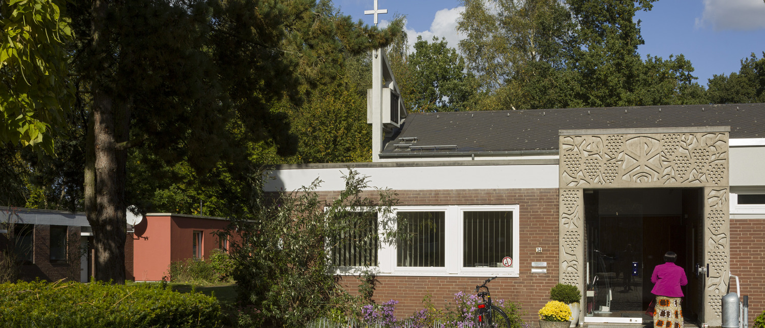 Ein modernes Gebäude mit einer kunstvoll verzierten Fassade und einem Kreuz auf dem Dach. Im Vordergrund steht eine Person in pinkem Oberteil, die zur Eingangstür schreitet. Bunte Blumen und ein Fahrrad sind ebenfalls sichtbar. Die Umgebung ist von Bäumen umgeben.