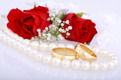 Auf dem Foto sieht man rote Rosen, Trauringe und eine Perlenkette