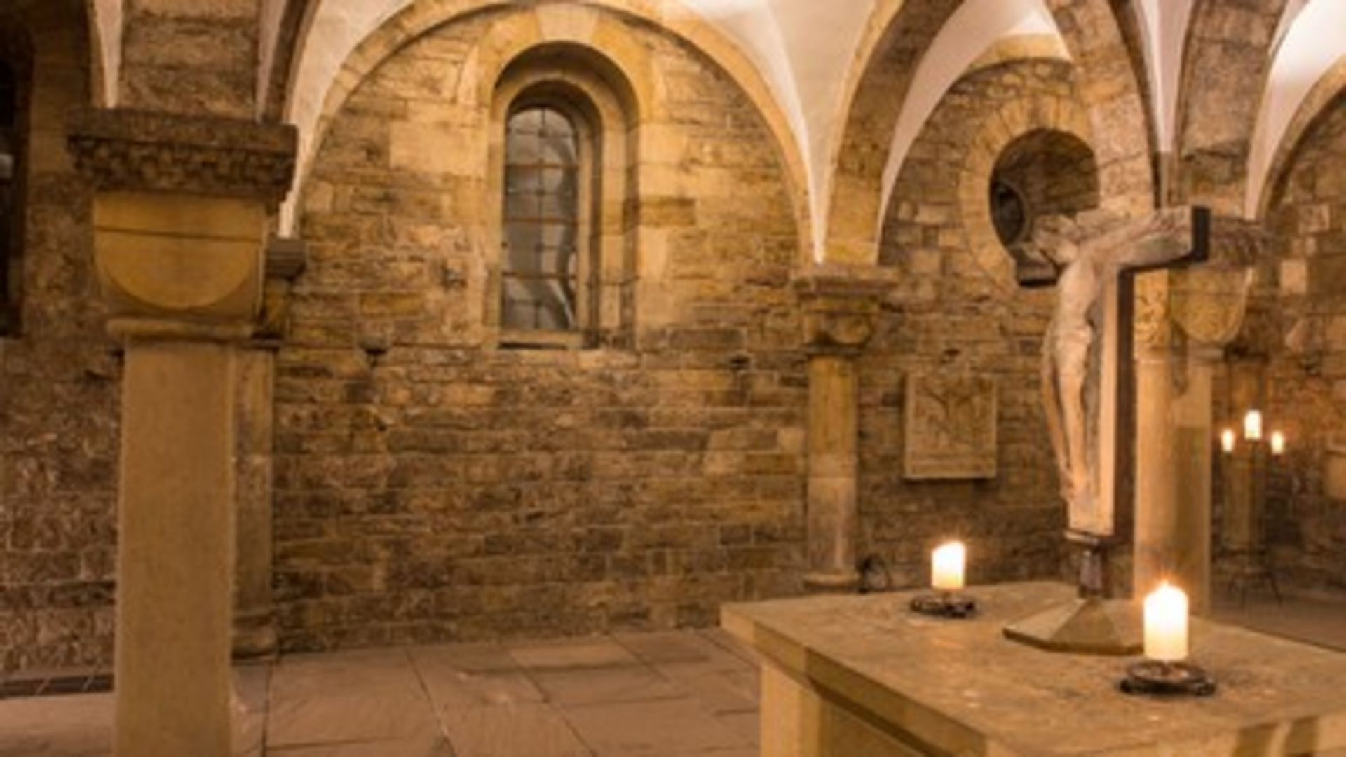 Der Raum zeigt eine historische Kirche mit Steinmauern und hohen, gewölbten Decken. In der Mitte steht ein Altar mit einer Kreuzigungsstatue und brennenden Kerzen, die eine ruhige, fromme Atmosphäre schaffen. Ein Fenster lässt gedämpftes Licht hinein.