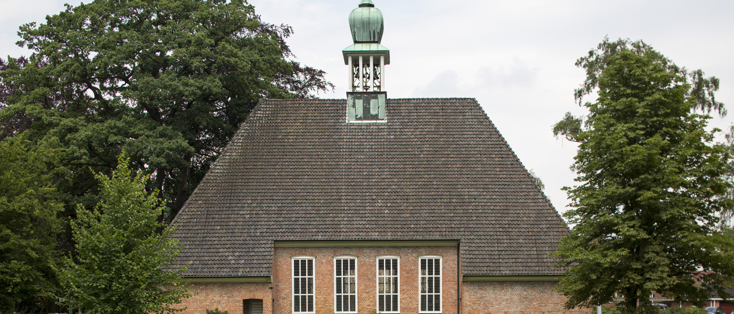 Eine rote Backsteinkirche mit einem hohen, spitzen Dach und einer kleinen Kupferkuppel. Vor der Kirche befinden sich grüne Wiesen und Bäume. Die Kirche hat große Fenster mit weißen Rahmen, die das Licht hereinlassen.