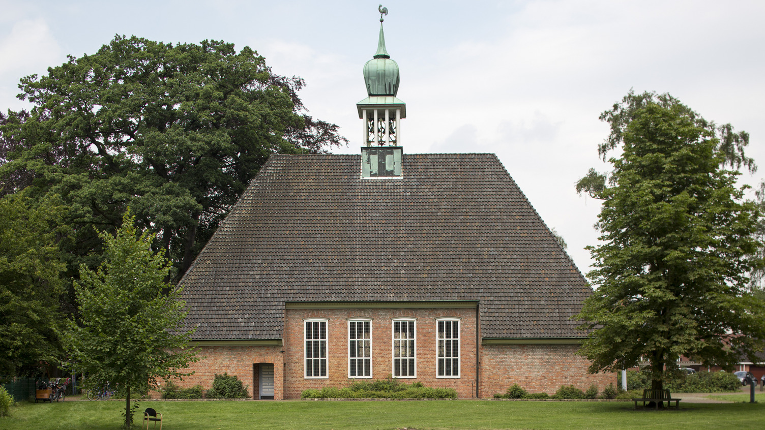 Eine rote Backsteinkirche mit einem hohen, spitzen Dach und einer kleinen Kupferkuppel. Vor der Kirche befinden sich grüne Wiesen und Bäume. Die Kirche hat große Fenster mit weißen Rahmen, die das Licht hereinlassen.