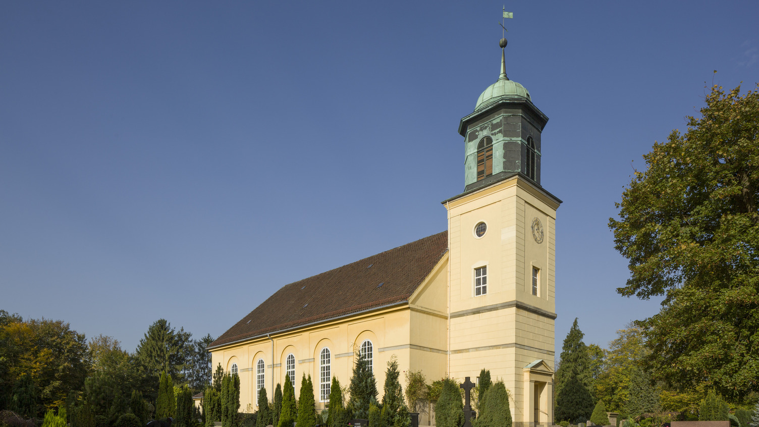 Eine gelbe Kirche mit einem markanten Turm steht umgeben von einem Friedhof mit Grabsteinen und bunten Blumen. Ein klarer blauer Himmel bildet den Hintergrund. Der Turm hat eine grüne Kuppel und Fenster.