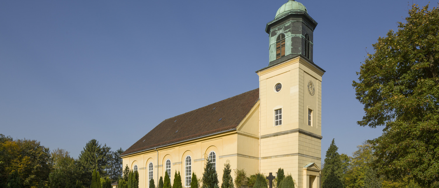 Eine gelbe Kirche mit einem markanten Turm steht umgeben von einem Friedhof mit Grabsteinen und bunten Blumen. Ein klarer blauer Himmel bildet den Hintergrund. Der Turm hat eine grüne Kuppel und Fenster.