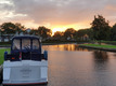 Man sieht ein Boot von hinten, welches am Ufer festgemacht ist. Im Hintergrund geht die Sonne über ein paar Häusern unter.