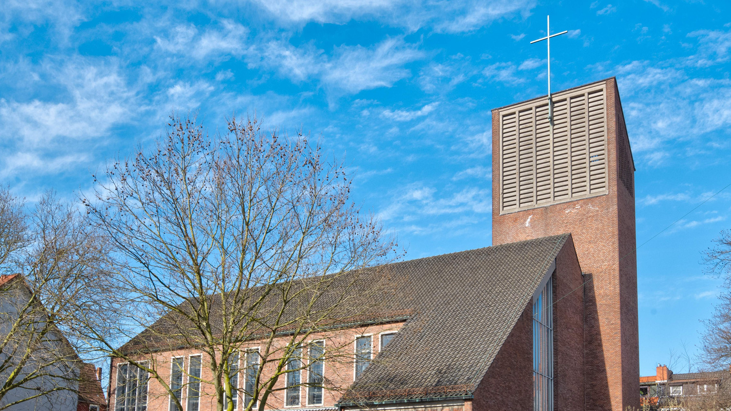 Eine moderne Kirche mit einem hohen, rechteckigen Turm und einem Kreuz an der Spitze. Die Fassade ist aus roten Ziegeln, mit großen Fenstern, die Tageslicht hereinlassen. Im Vordergrund sind kahle Bäume sichtbar, vor einem klaren blauen Himmel.