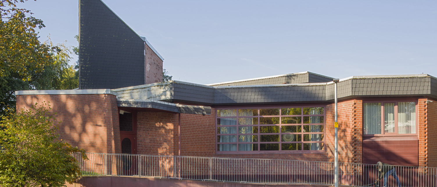 Ein modernes Gebäude mit roter Backsteinfassade und einem hohen, schrägen Dach. An der Spitze des Daches befindet sich ein metallisches Kreuz. Große Fenster lassen viel Tageslicht herein, und ein Weg führt zum Eingang. Bäume und ein gepflasterter Gehweg umgeben das Gebäude.