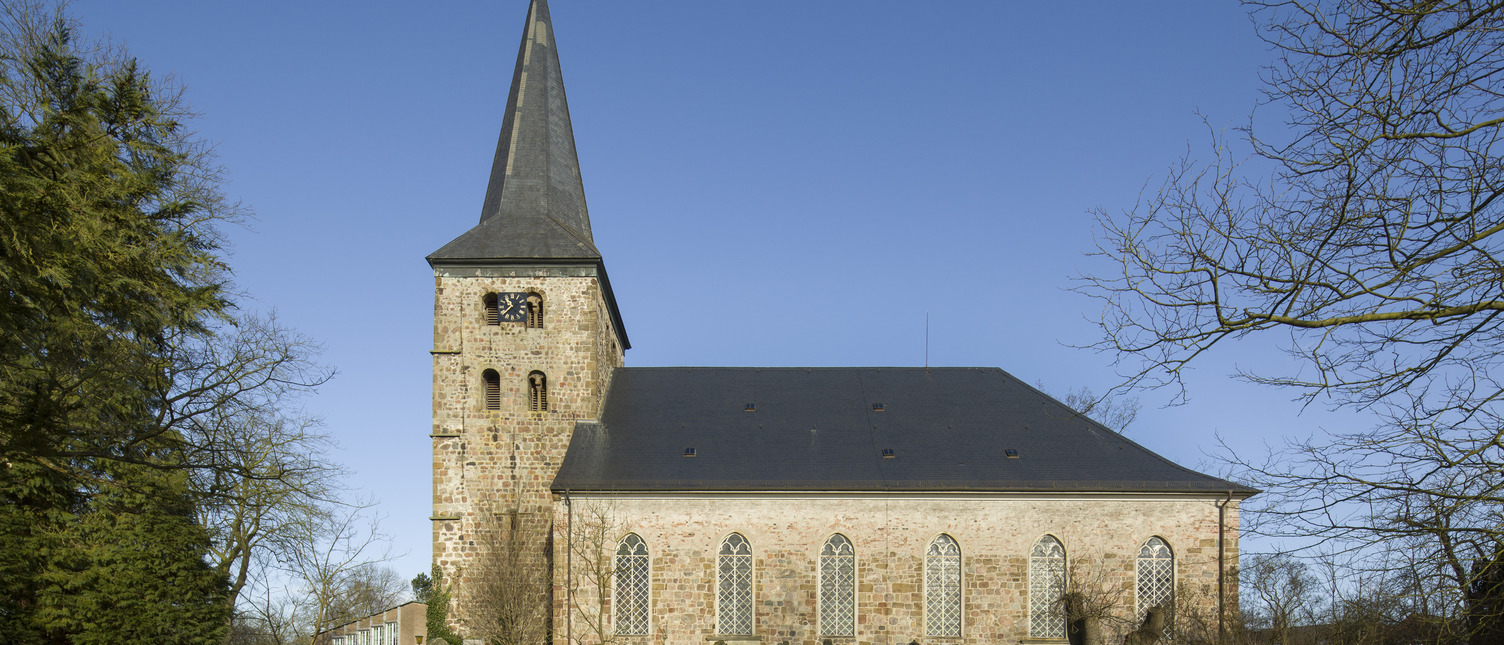Die Bildbeschreibung zeigt eine Kirche mit einem hohen, spitzen Turm und charakteristischen Fenstern. Der Gebäudekörper ist aus helleren Ziegelsteinen, umgeben von einem ruhigen, grünen Rasen und einigen Grabsteinen im Vordergrund. Der Himmel ist klar und blau.