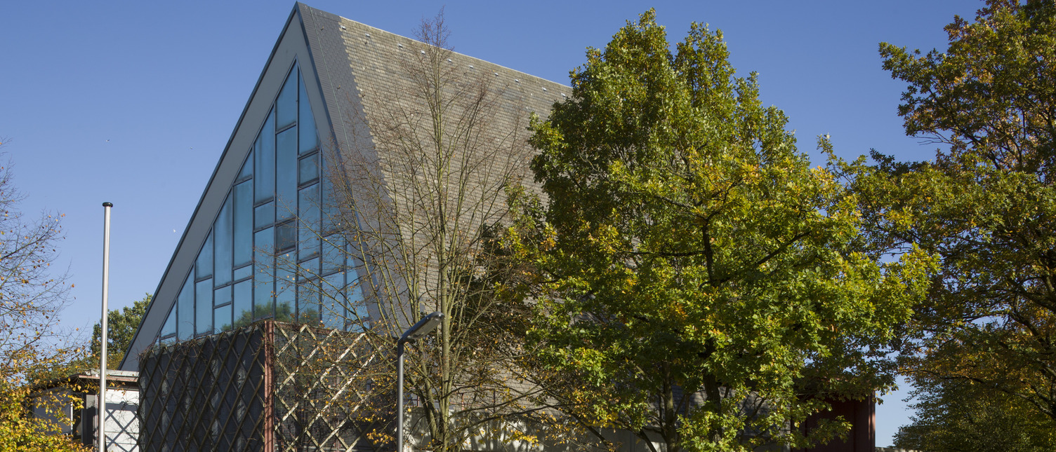 Ein modernes Gebäude mit schräg gestuftem Dach und großen Glasfenstern, umgeben von Bäumen. Die Fassade ist teilweise mit einer dekorativen Gitterstruktur verkleidet. Der Himmel ist klar und blau.