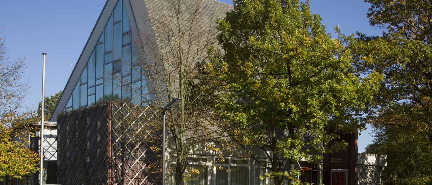 Ein modernes Gebäude mit schräg gestuftem Dach und großen Glasfenstern, umgeben von Bäumen. Die Fassade ist teilweise mit einer dekorativen Gitterstruktur verkleidet. Der Himmel ist klar und blau.