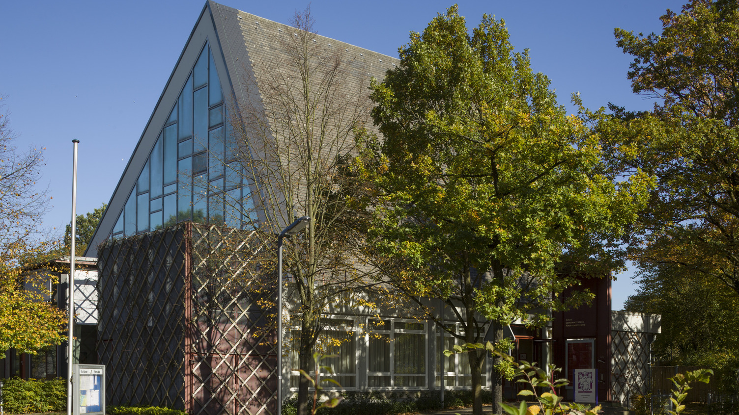 Ein modernes Gebäude mit schräg gestuftem Dach und großen Glasfenstern, umgeben von Bäumen. Die Fassade ist teilweise mit einer dekorativen Gitterstruktur verkleidet. Der Himmel ist klar und blau.