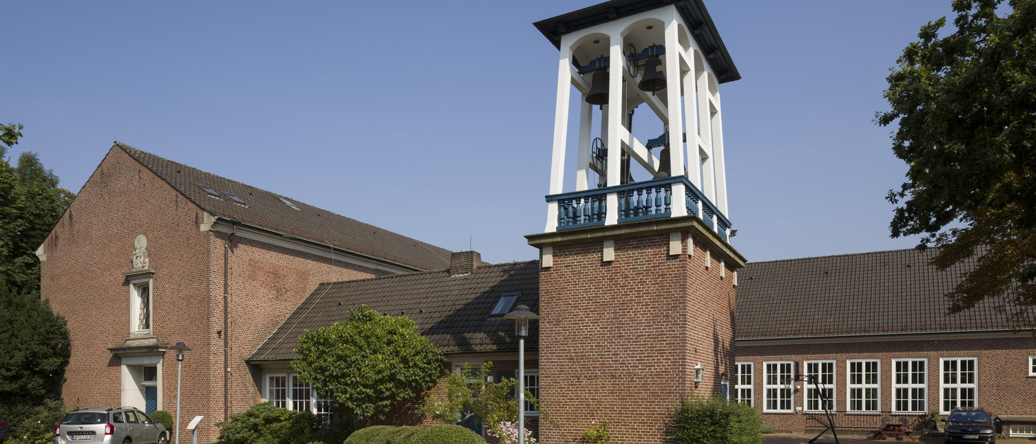 Ein rotes Backsteingebäude mit einem schlichten, weißen Glockenturm und mehreren Fenstern. Umgeben von grünen Pflanzen und einem gepflasterten Vorplatz. Der Himmel ist blau und wolkenlos.