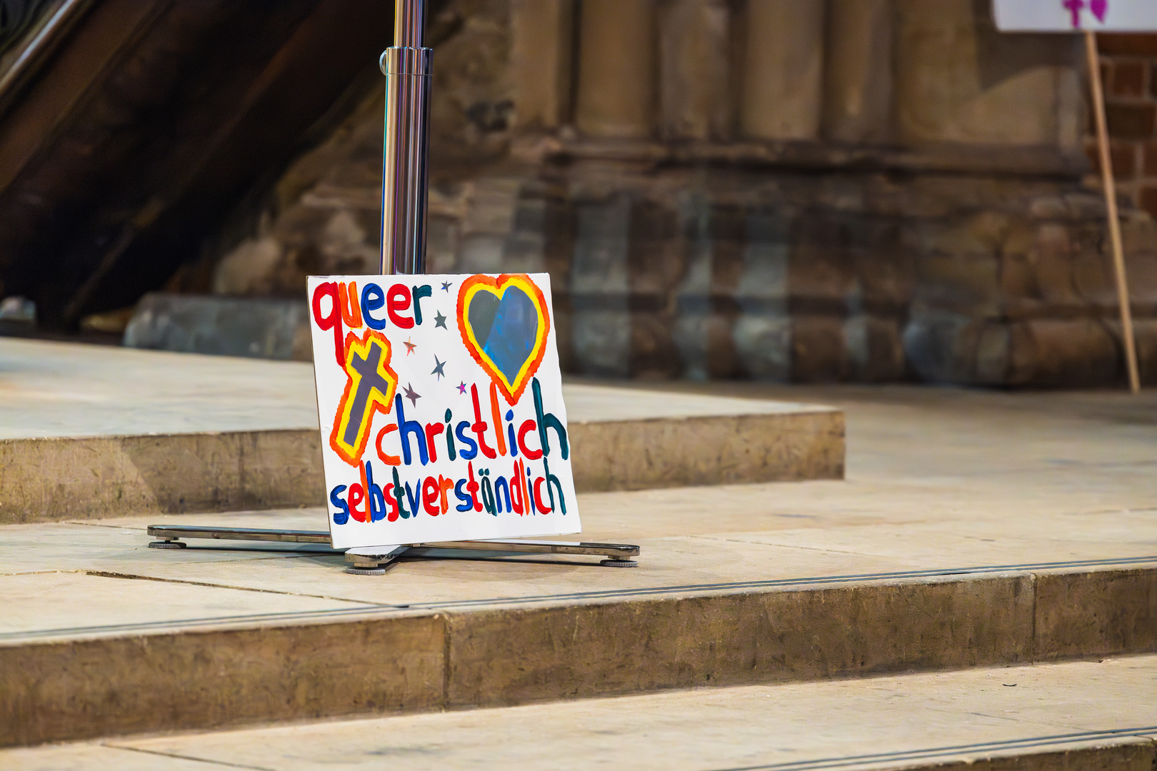 Ein Pappschild lehnt an den Stufen, die zum Altar der Kirche führen. Auf dem Schild steht: "Queer - christlich - selbstverständlich". Außerdem sind ein großes Kreuz und ein großes Herz und ein paar kleine Sterne aus glänzender Folie auf dem Schild zu sehen.