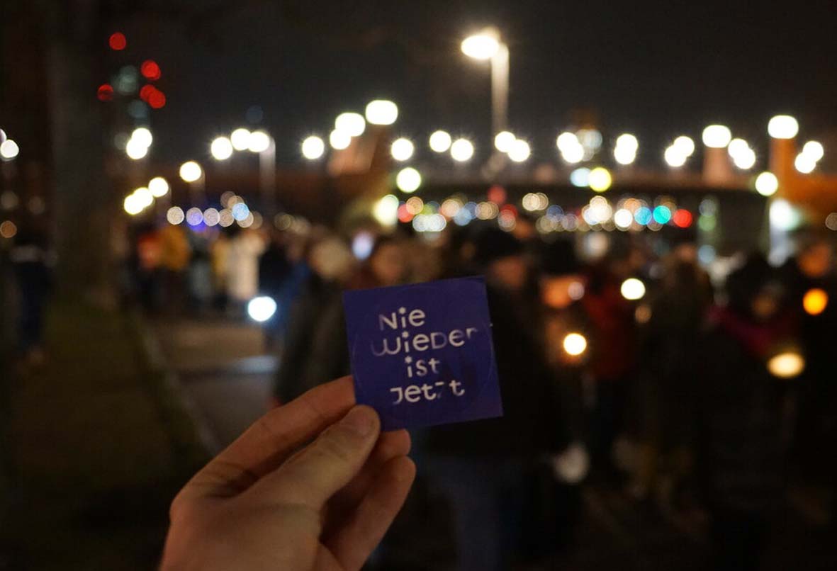 Das Foto zeigt eine Lichterketten-Demo. Im Vordergrund hält eine Hand ein Schild mit der Aufschrift "Nie wieder ist jetzt" hoch